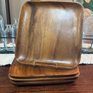 Pode incluir: Conjunto de quatro travessas quadradas de madeira. As travessas são feitas de madeira de tons quentes com grão visível. Uma pega de metal cor de latão está presa à travessa superior. As travessas estão empilhadas umas sobre as outras.