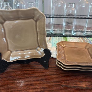 Può includere: Un set di piatti quadrati in ceramica color beige con bordi smerlati. Un piatto è esposto su un supporto nero, mentre una pila di quattro piatti poggia su una superficie di legno. I piatti hanno una finitura lucida.