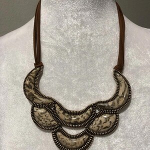 May include: A statement necklace featuring a brown suede cord and multiple crescent-shaped pendants. The pendants have a speckled, off-white and brown pattern, framed by a metallic zipper-like trim. The necklace is displayed on a mannequin.