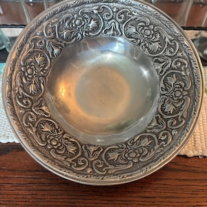 May include: A silver-colored, antique bowl with an ornate floral and scrollwork design around the rim. The bowl has a shallow, rounded center and is likely made of metal. The intricate details suggest a vintage or handcrafted piece.