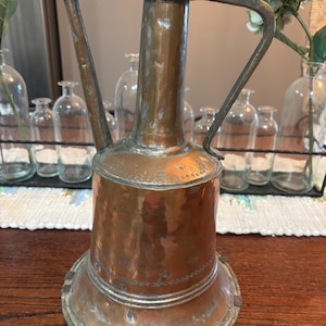 May include: An antique copper pitcher with a handle and spout. The pitcher has a wide base and a cylindrical body, with a decorative band around the middle. The metal has a tarnished, aged appearance, suggesting it is vintage. The pitcher is likely used for decorative purposes.