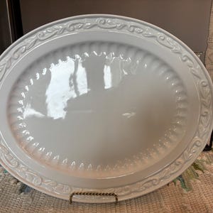 Puede incluir: Una bandeja de servir ovalada de cerámica blanca con un centro liso y brillante, rodeada por un borde decorativo en relieve con un patrón de volutas y hojas, y una textura estriada sutil a lo largo del borde interior.