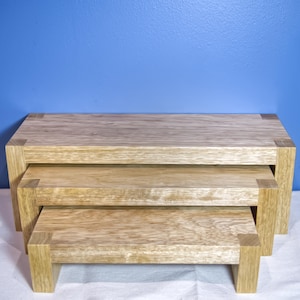 May include: A set of three wooden nesting tables. The tables are rectangular with a light wood grain finish. The largest table is at the top, with the other two nested below. The tables have square corner accents and are set against a blue background.