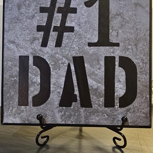 May include: Square decorative tile with the words "#1 DAD" in bold, black stencil lettering. The tile has a textured, grey background and is displayed on a black metal stand. A great gift for Father's Day.
