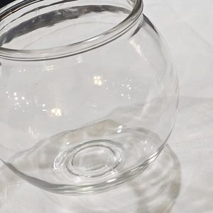 Può includere: Una ciotola in vetro trasparente di forma sferica con un'ampia apertura superiore e una base circolare spessa e rinforzata. Il recipiente presenta pareti lisce e curve che si restringono verso il bordo, creando una forma classica.