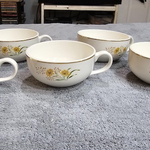 May include: A set of four cream-colored ceramic soup bowls with handles, each featuring a delicate floral pattern of yellow flowers and green stems on the exterior, finished with a thin metallic gold rim around the top edge of each bowl.