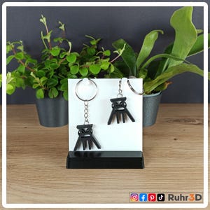 Könnte beinhalten: Zwei schwarze Schlüsselanhänger mit einem stilisierten Design einer Industriestruktur. Die Schlüsselanhänger werden auf einem weißen Hintergrund mit schwarzem Sockel präsentiert. Der Hintergrund umfasst Topfpflanzen und den Text "Ruhr3D" mit Social-Media-Symbolen.