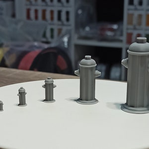 Op de afbeelding: Vijf grijze plastic brandkranen in verschillende maten, gerangschikt in een rij van de kleinste tot de grootste op een wit rond platform.