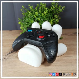 Könnte beinhalten: Ein schwarzer kabelloser Gamecontroller mit blauen und roten Knöpfen liegt auf einem weißen, abstrakt geformten Ständer. Der Controller hat einen strukturierten Griff und ein Steuerkreuz. Im Hintergrund befinden sich eine grüne Pflanze und eine Holzoberfläche.