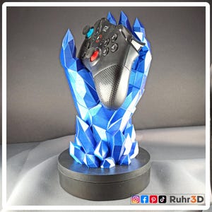 Könnte beinhalten: Ein schwarzer Gamecontroller ruht in einem blauen, kristallförmigen Halter. Der Halter besteht aus blauem Kunststoff und steht auf einem schwarzen Sockel. Der Controller hat rote, blaue und schwarze Tasten. Das Design ist futuristisch und dekorativ.