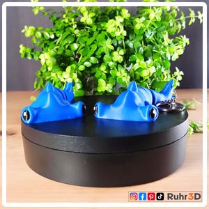 Könnte beinhalten: Zwei blaue Hammerhai-Figuren mit schwarzen Augen und einem Schlüsselanhänger. Die Figuren stehen auf einer schwarzen Oberfläche, im Hintergrund eine grüne Pflanze. Die Figuren sind 3D-gedruckt.