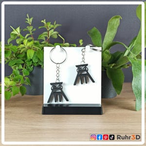 May include: Two black keychains with a stylized mining tower design are displayed on a white stand. The keychains hang from silver chains and rings. The stand is on a wooden surface, with green plants in the background. The logo "Ruhr3D" is visible.