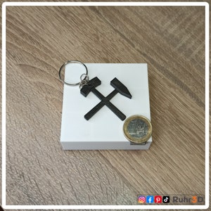 Könnte beinhalten: Ein schwarzer 3D-gedruckter Schlüsselanhänger mit einem Design aus gekreuztem Pickel und Hammer. Der Schlüsselanhänger befindet sich auf einem weißen Hintergrund mit einer Euro-Münze als Größenvergleich.