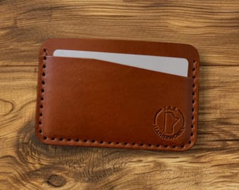 May include: A brown leather card holder with a stitched border and a logo. The wallet has a pocket with a white card partially visible. The card holder is on a wooden surface.