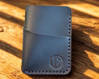 May include: A dark blue leather card wallet with a curved pocket and stitched edges. The wallet has a circular embossed logo on the front. The wallet is sitting on a wooden surface.
