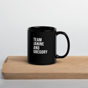 Puede incluir: Taza de café de cerámica negra con texto blanco que dice "TEAM JANINE AND GREGORY".