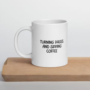 Può includere: Tazza da caffè in ceramica bianca con testo nero che recita "Turning pages and sipping coffee".