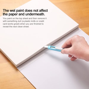 May include: A white pad of paper with a blue utility knife cutting through the top sheet. The text on the pad reads: "The wet paint does not affect the paper and underneath. You paint on the top sheet and then remove it with something dull (a plastic knife or credit card works great) when you are finished to reveal the next clean sheet."