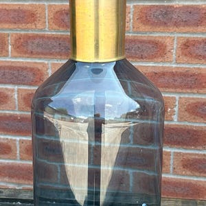 May include: A tall, cylindrical glass decanter featuring a deep blue-grey tint. The vessel has a smooth, straight-sided body, a polished gold-toned metal neck, and a large, spherical glass stopper in a matching blue-grey hue.