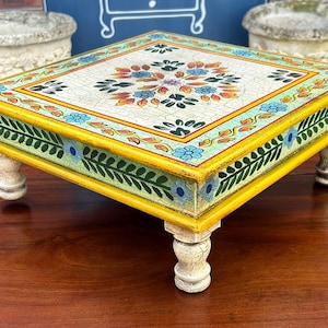 May include: A square wooden pedestal stand featuring a hand-painted floral motif with a crackled white finish on the top surface. The sides are decorated with green leaf patterns and blue flowers, framed by a bright yellow border on four carved legs.