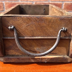 May include: A rectangular wooden box with a curved metal handle. The box has a dark brown finish and rounded edges. The metal handle is dark grey and is attached to the front of the box. The box is likely for storage.