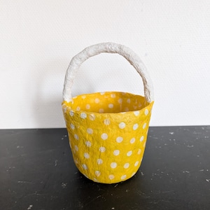 Può includere: Un piccolo cestino giallo fatto a mano con pois bianchi. Il cestino ha un manico bianco ed è probabilmente fatto di carta o materiale simile. Il cestino è appoggiato su una superficie nera.