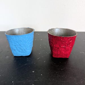 Peut inclure: Deux petits gobelets en papier texturé, l'un bleu vif, l'autre rouge vif. Les deux ont un intérieur argenté. Les gobelets sont posés sur une surface sombre.