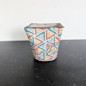 Peut inclure: Un petit pot de fleurs carré avec un motif géométrique coloré de triangles bleus, rouges et jaunes. Le bord est souligné en noir. Le pot est posé sur une surface sombre.