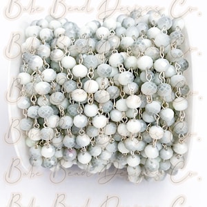 May include: A close-up of a silver chain with white and gray faceted glass beads. The chain is coiled and the beads are visible from all angles.