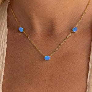May include: A delicate gold necklace featuring three oval, light blue gemstones. The gemstones are evenly spaced along the fine chain. The necklace is worn against a neutral-toned top.
