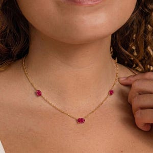 May include: A delicate gold necklace featuring three oval-shaped, vibrant red gemstones. The necklace is worn around the neck, showcasing the gemstones' rich color and the chain's subtle shine. The design is simple yet elegant, suitable for various occasions.