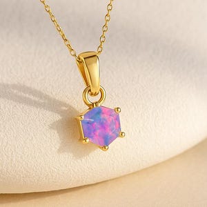 May include: A gold-colored necklace with a hexagonal pendant. The pendant features a pink and blue opal stone. The chain and pendant setting are a bright gold color. The necklace is displayed on a cream-colored surface.