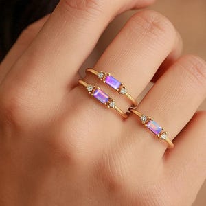 Könnte beinhalten: Drei goldene Ringe mit rechteckigen, irisierenden lila-blauen Steinen und kleinen Diamantakzenten. Die Ringe werden auf einer Hand präsentiert, um ihr zartes Design und ihr elegantes Aussehen hervorzuheben. Der Schmuck ist ideal, um einen Hauch von Raffinesse zu verleihen.