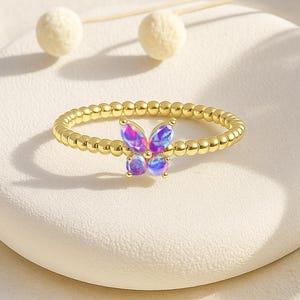 Puede incluir: Un anillo dorado con un dise&ntilde;o de mariposa con alas iridiscentes moradas y azules. La banda del anillo est&aacute; adornada con peque&ntilde;as cuentas espaciadas uniformemente. La mariposa es el punto focal, agregando un toque de elegancia inspirada en la naturaleza.