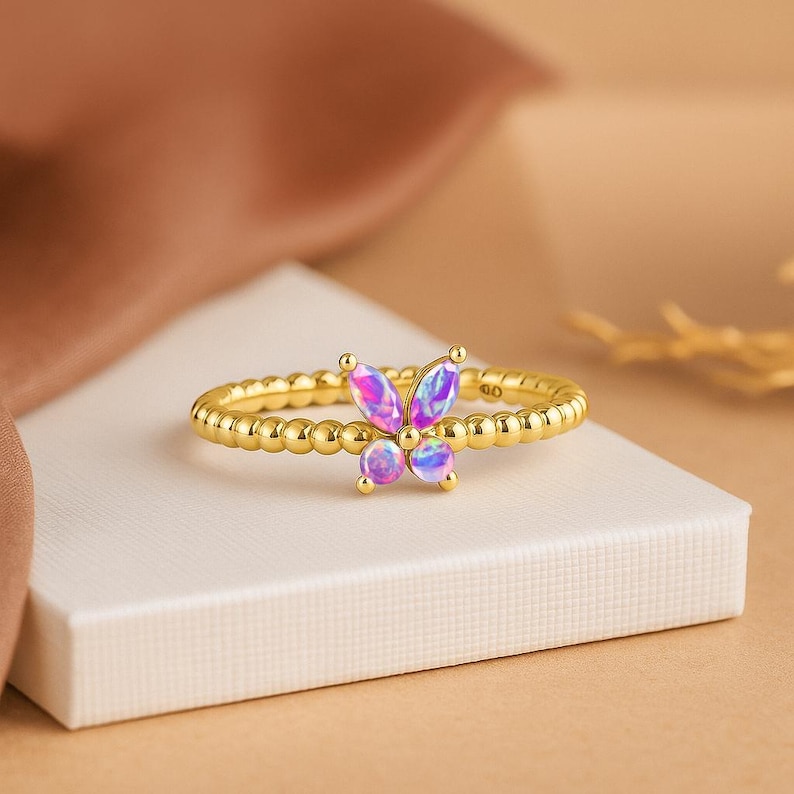 Puede incluir: Un anillo dorado con un dise&ntilde;o de mariposa. La mariposa tiene piedras iridiscentes rosas y moradas. La banda del anillo tiene una textura con peque&ntilde;as cuentas redondas. El anillo se muestra sobre una superficie blanca.