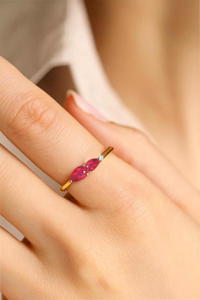 K&ouml;nnte beinhalten: Ein goldener Ring mit zwei markant roten, marquisef&ouml;rmigen Edelsteinen, die nebeneinander gesetzt sind. Zwischen den roten Steinen befindet sich ein kleiner, runder, klarer Edelstein. Der Ring befindet sich an einem Finger.