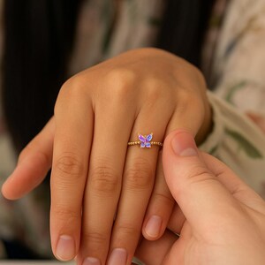 Puede incluir: Un anillo dorado con una peque&ntilde;a mariposa con alas iridiscentes moradas y azules. El anillo est&aacute; en un dedo, con otra mano toc&aacute;ndolo suavemente. La banda del anillo tiene un dise&ntilde;o de cuentas.