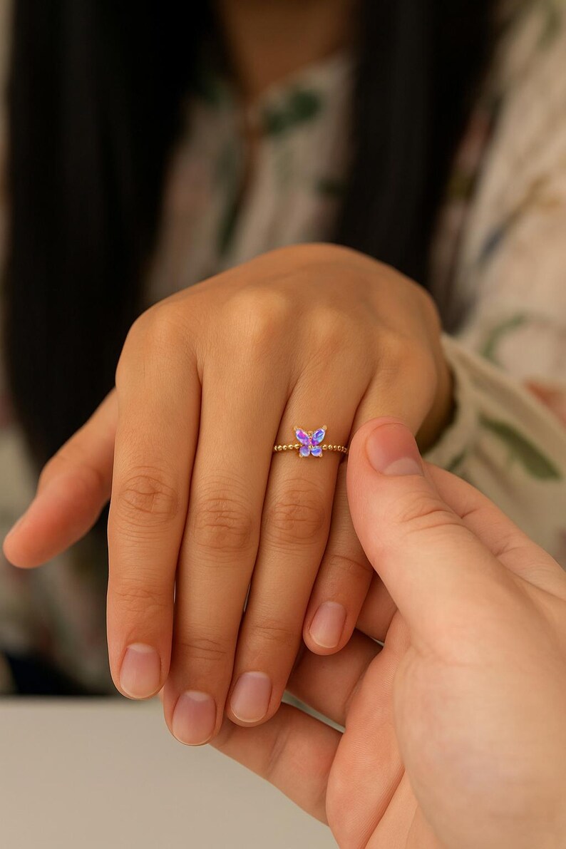 Puede incluir: Un anillo dorado con una peque&ntilde;a mariposa con alas iridiscentes moradas y azules. El anillo est&aacute; en un dedo, con otra mano toc&aacute;ndolo suavemente. La banda del anillo tiene un dise&ntilde;o de cuentas.