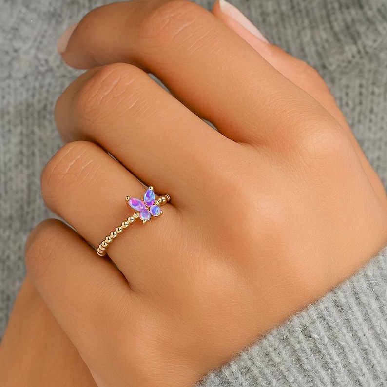 Puede incluir: Un anillo dorado con un dise&ntilde;o de mariposa con acentos iridiscentes morados y azules. La banda del anillo tiene una textura de cuentas. La mariposa es el punto focal, agregando un toque de fantas&iacute;a a la joyer&iacute;a.