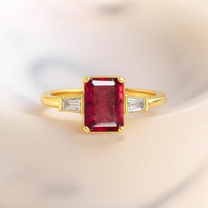 Peut inclure: Une bague en or ornée d'une grande pierre rouge foncé rectangulaire au centre. Deux petites pierres claires rectangulaires flanquent la pierre principale. La bague présente un design classique et élégant.