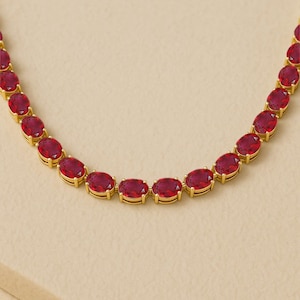 May include: A gold necklace with a series of oval-cut, deep red gemstones. The gemstones are set in individual gold bezels, creating a continuous line of vibrant colour. The necklace is displayed against a neutral background.
