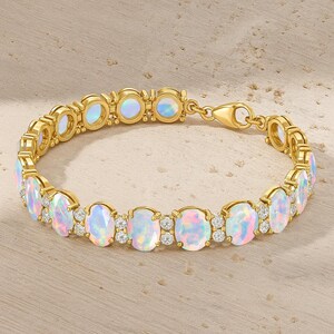 May include: A gold bracelet with oval opals and small, clear gemstones. The bracelet has a clasp closure. The opals display pink, blue, and white hues. The bracelet is set on a light-colored surface. The bracelet is approximately 19 cm in length.