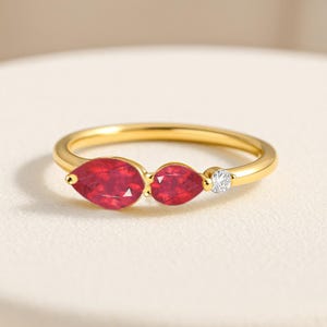 Könnte beinhalten: Ein goldener Ring mit drei Edelsteinen. Zwei tropfenförmige, tiefrote Rubine sind nebeneinander gesetzt, mit einem kleinen, runden, klaren Diamanten rechts. Das Ringband ist aus poliertem Gold.