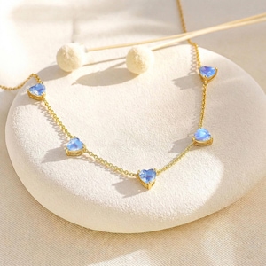 May include: A delicate gold necklace featuring five heart-shaped, light blue gemstones. The necklace is displayed on a white, textured surface, with a soft, neutral background. The chain is fine and the gemstones are evenly spaced.