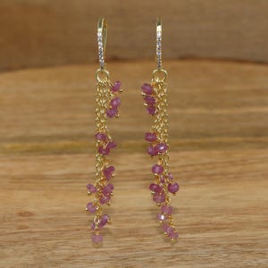 May include: A pair of long gold dangle earrings featuring multiple delicate gold chains adorned with small, faceted pink gemstone beads. The top of each earring has a curved gold hook encrusted with tiny clear stones for a secure fit.