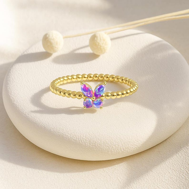 Puede incluir: Un anillo dorado con un dise&ntilde;o de mariposa con alas iridiscentes moradas y azules. La banda del anillo est&aacute; adornada con peque&ntilde;as cuentas espaciadas uniformemente. La mariposa es el punto focal, agregando un toque de elegancia inspirada en la naturaleza.