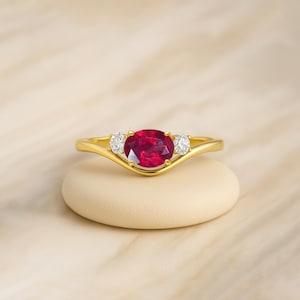 Peut inclure: Une bague en or ornée d'un grand rubis ovale et de deux petites pierres précieuses claires. La bague est posée sur une surface arrondie de couleur claire, sur un fond doux et neutre. Le design est élégant.