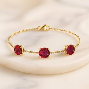 May include: A gold bracelet with three large, round, deep red gemstones. The gemstones are set in square gold settings and linked by a delicate gold chain. The bracelet is displayed on a white surface.
