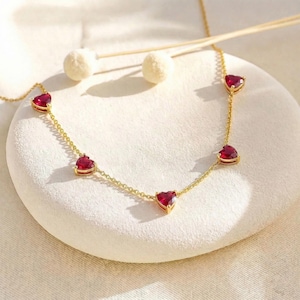 Könnte beinhalten: Eine zarte goldene Halskette mit herzförmigen roten Edelsteinen. Die Halskette hat eine feine Kette mit gleichmäßig verteilten Herzanhängern. Die Edelsteine sind leuchtend rot und in Goldfassungen gefasst. Die Halskette wird auf einer weißen Oberfläche präsentiert.