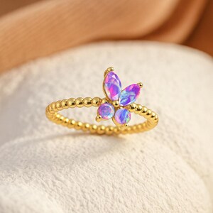Puede incluir: Anillo dorado con dise&ntilde;o de mariposa con piedras preciosas iridiscentes moradas y azules. La banda del anillo tiene una textura de cuentas. La mariposa tiene tres piedras redondas y tres piedras en forma de marquesa.